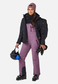 Skikleding met een zwarte pufferjack, paarse skibroek en zwarte skischoenen. Accessoires zijn een roze muts en skibril.