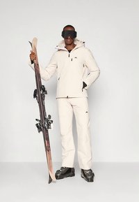 Άνδρας φορώντας μπεζ σκι jacket, παντελόνι, μαύρα μποτάκια σκι και γυαλιά σκι κρατάει σκι κατακόρυφα σε ουδέτερο φόντο.