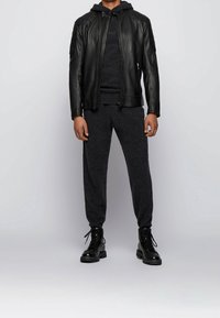 Černá kožená bunda s prošívanými detaily na ramenou, šedá pletená mikina s kapucí, odpovídající šedé tepláky a černé šněrovací boty.