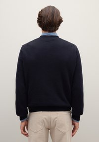 Uomo con capelli castani che indossa un maglione blu navy sopra una camicia blu e pantaloni beige, in piedi di fronte a una parete bianco sporco.