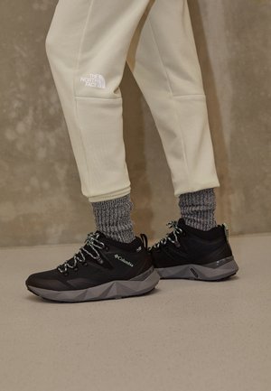 Persona che indossa pantaloni beige North Face, calzini grigi lavorati a maglia e scarpe da trekking nere Columbia, in piedi su un pavimento chiaro.