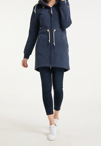 Femme portant un manteau à capuche bleu marine zippé avec des cordons blancs, un jean skinny foncé et des baskets blanches, marchant sur un fond uni.