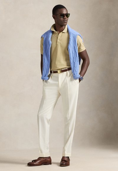 Άνδρας με γυαλιά ηλίου, μπεζ πόλο μπλούζα, λευκό παντελόνι, καφέ μοκασίνια, μπλε πουλόβερ πάνω στους ώμους, στέκεται με τα χέρια στις τσέπες.