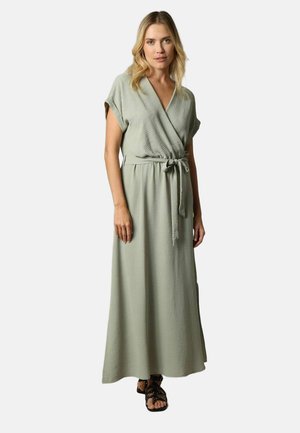 Femme portant une robe maxi portefeuille à manches courtes vert clair avec une ceinture à la taille et des sandales plates noires à brides, se tenant devant un fond neutre.