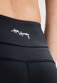 Zbliżenie na talię osoby w czarnych legginsach z złożonym pasem i białym logo "Hey Honey" z tyłu.