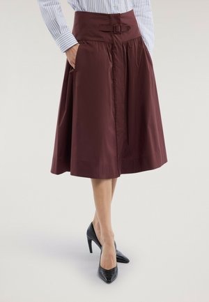 Frau trägt einen midi-langen bordeauxroten Rock mit Schnallendetail und Taschen, kombiniert mit gestreiftem Hemd und schwarzen High-Heels.