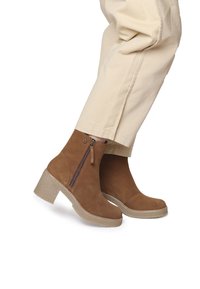 Stivali alla caviglia in suede marrone con cerniera laterale, suola texturizzata di colore chiaro e tacco chunky per stabilità. I pantaloni sono beige.