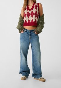 Cropped svetr s červeným a krémovým argyle vzorem navrstvený na olivově zelené bundě; doplněný volnými modrými denimovými džíny a tanovými teniskami.