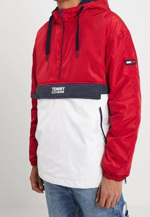 Czerwono-biała wiatrówka Tommy Jeans z granatowymi troczkami i szarym obszyciem, noszona przez osobę w niebieskich dżinsach.