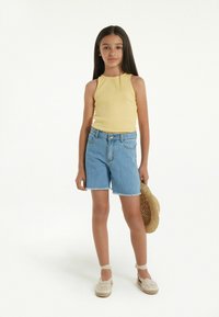 Débardeur jaune à côtes avec un col rond, associé à un short en denim bleu clair avec un ourlet effiloché. Le modèle tient un chapeau en paille.