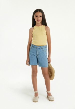 Débardeur jaune à côtes avec un col rond, associé à un short en denim bleu clair avec un ourlet effiloché. Le modèle tient un chapeau en paille.