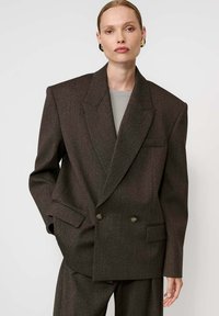 Modell oversized, sötétbarna, dupla gombos blézerben, széles hajtókákkal, hozzá illő nadrágban, szürke felsőben és arany karikafülbevalóban, fehér háttér előtt.