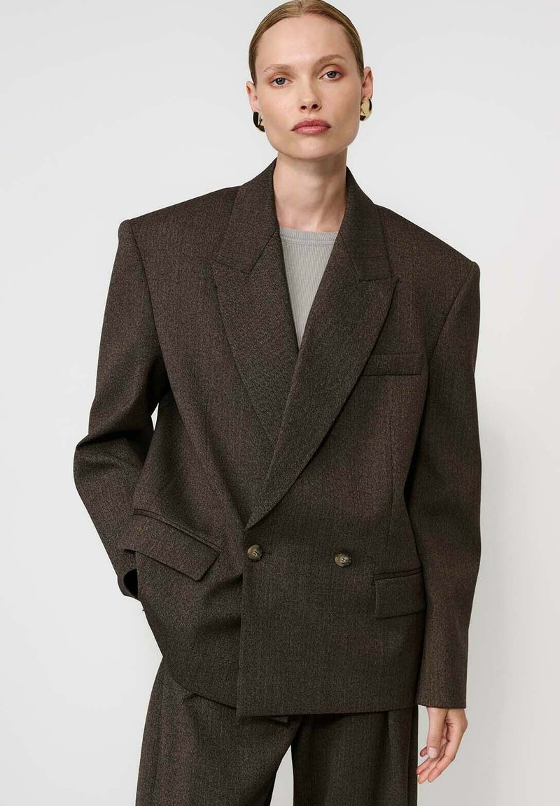 Modell oversized, sötétbarna, dupla gombos blézerben, széles hajtókákkal, hozzá illő nadrágban, szürke felsőben és arany karikafülbevalóban, fehér háttér előtt.