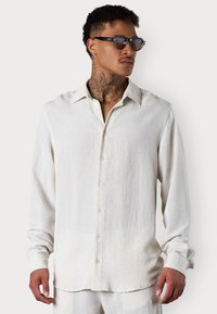 Helles beige Hemd mit Knöpfen aus strukturiertem Stoff, mit Kragen, langen Ärmeln und Vorderknöpfen. Keine Muster oder Akzente vorhanden.