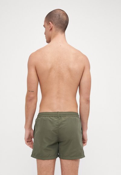 Shorts de bain verts avec une taille élastique et des poches latérales, fabriqués dans un tissu lisse et léger. Vue de l'arrière d'une silhouette masculine.