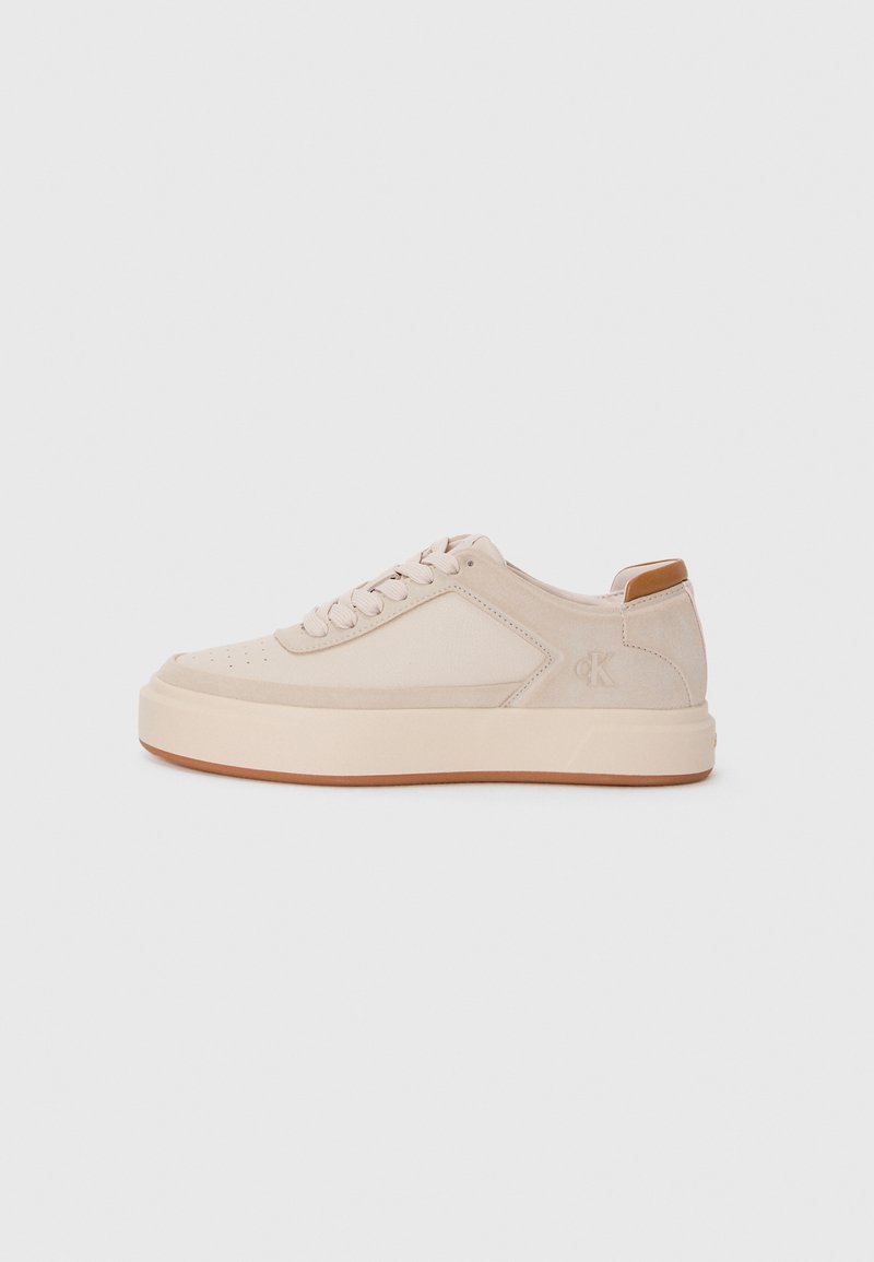 Beige Sneakers mit Wildleder-Oberfläche, runder Zehenpartie, flacher Gummisohle, Schnürdesign und einem kleinen, braunen Akzent an der Ferse mit einem Logo.