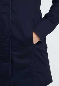 Marineblauwe katoenen jas met een rechte snit, zijzakken en contrasterende stiksels. Voorzien van een label aan de lagere zijnaden.