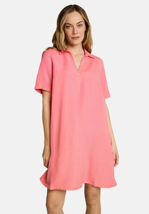 Mujer con vestido rosa holgado de manga corta, con cuello y escote en V, de pie frente a un fondo blanco.