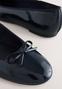 Ballerines noires brillantes avec un petit nœud à l'avant et un bord élastique autour de l'ouverture, présentées sur un fond neutre.