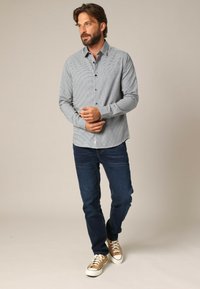 Man met een baard in een grijs, klein-geruit button-up shirt, donkerblauwe jeans en beige hoge sneakers, staand tegen een beige achtergrond.