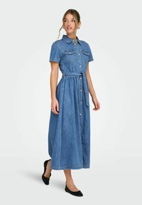 Femme debout, portant une robe midi en denim bleue boutonnée avec des manches courtes, une ceinture à la taille, et des chaussures plates noires, regardant sur le côté.