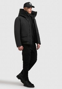 Juodas paminkštintas švarkas su aukštu apykakle, priekinėmis kišenėmis, dėvimas virš juodos kapšos, derinamas su juodomis kariškų stilių kelnėmis ir tvirtais juodais batais.