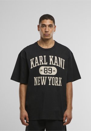 Junger Mann mit kurzen dunklen Haaren und Creolen, trägt ein lockeres schwarzes T-Shirt mit dem Text "KARL KANI 89 NEW YORK".