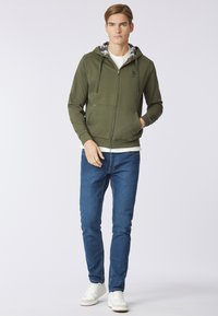 Felpa verde con zip e cappuccio a quadri, abbinata a jeans slim-fit blu e sneakers bianche. Tessuto morbido e design casual.