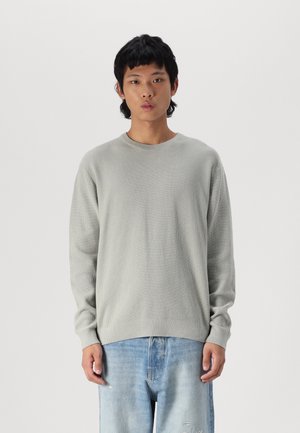 Jeune homme debout, face à l'avant, portant un pull en tricot gris clair et un jean délavé bleu clair, sur un fond uni.
