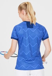 Blå atletisk skjorte med et geometrisk mønster med chevroner, korte ærmer og en ensfarvet blå krave. Stoffet virker let og åndbart.