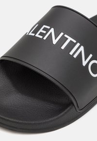 Black slide sandal made of smooth rubber with a wide strap featuring the white text "VALENTINO." Text is bold and centered.