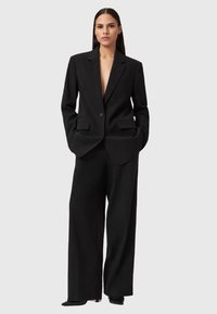Vrouw met lang, donker haar, gekleed in een oversized zwarte blazer en wijde broek, staande met haar handen in de zakken op een grijze achtergrond.