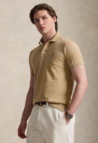 Young man with short brown hair wearing a beige polo shirt, white trousers, and a wristwatch, standing against a plain background.
