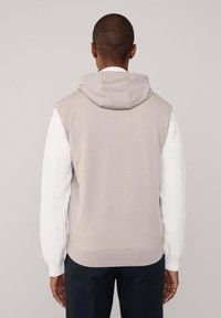Hoodie senza maniche di un taupe chiaro con finitura texturizzata e cappuccio, abbinato a una maglietta a maniche lunghe bianca a costine sotto, su uno sfondo neutro.