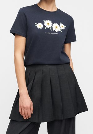 Granatowy t-shirt z białymi stokrotkami i tekstem "Live life in full bloom," zestawiony z czarną plisowaną spódnicą noszoną na ciemnych spodniach.