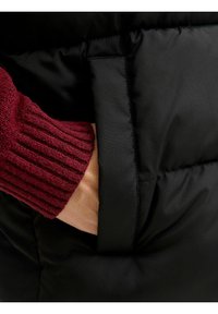 Chaleco acolchado negro con un bolsillo lateral visible y una manga acanalada burdeos asomando. Tela suave con un acabado mate.