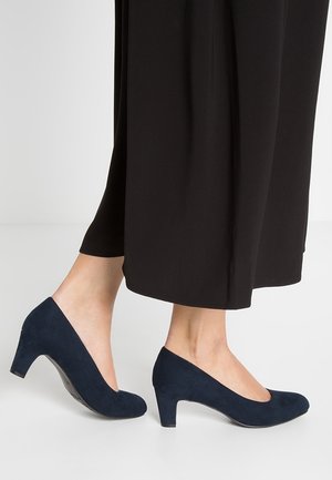 Escarpins en daim bleu marine à talons hauts avec un bout arrondi et un petit talon bloc, portés avec un pantalon noir à jambes larges. Texture lisse, design simple.