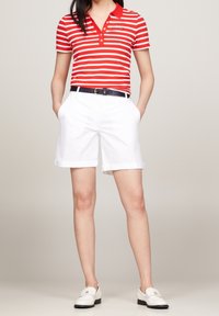 Maglietta polo a righe rosse e bianche con colletto e bottoni, abbinata a pantaloni corti bianchi fino al ginocchio e una cintura nera. Mocassini bianchi visibili.