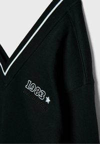Vêtement noir à col en V avec bordure blanche et texte brodé "1983" et "Manhattan" avec une petite étoile sur la zone de la poitrine.