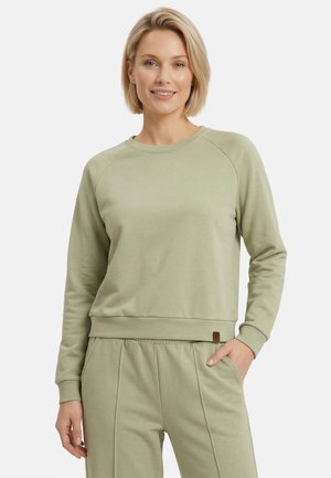 Vrouw met kort blond haar die een lichtgroene longsleeve trui en bijpassende broek draagt, glimlachend met één hand in de zak.