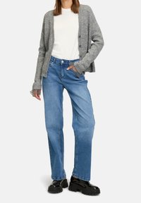 Szary dzianinowy kardigan na białej koszulce, zestawiony z niebieskimi szerokimi spodniami jeansowymi i czarnymi butami na masywnej podeszwie. Prosty krój i luźne dopasowanie.