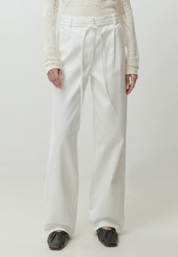 Witte, hoge taille, wijde broek met een trekkoord, gemaakt van een gestructureerde stof. Gecombineerd met zwarte vierkante schoenen.