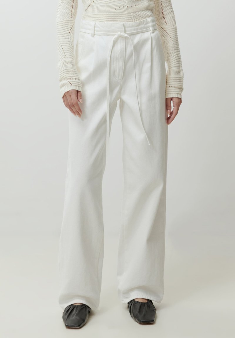 Witte, hoge taille, wijde broek met een trekkoord, gemaakt van een gestructureerde stof. Gecombineerd met zwarte vierkante schoenen.