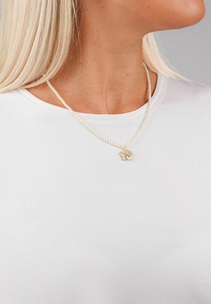 Pärlhalsband med en blommig hänge, som har en delikat design och mjuk textur. Bärs över en vit topp med rund halsringning.