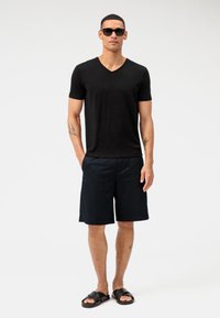 T-shirt nero a maniche corte e scollo a V, pantaloni corti blu navy con tasche e sandali neri. Persona che indossa occhiali da sole si trova di fronte a uno sfondo semplice.