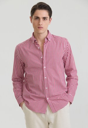 Junger Mann mit kurzen dunklen Haaren, der ein rot-weiß kariertes Hemd mit Knöpfen und hellbeige Hosen trägt, steht mit den Händen in den Taschen.