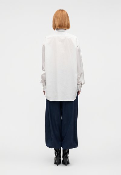 Λευκό πουκάμισο με μακριά μανίκια και γιακά, συνδυασμένο με ναυτικού μπλε παντελόνι φαρδιάς γραμμής με λεπτές ρίγες. Το σύνολο ολοκληρώνεται με μαύρες μπότες με ψηλό τακούνι.