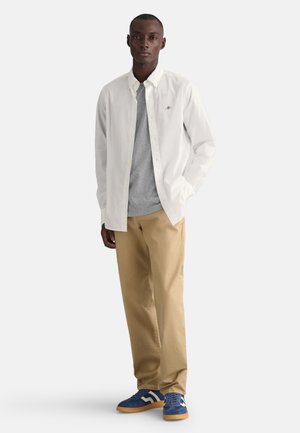 Witte overhemd over een grijs T-shirt, beige broek en blauwe suède sneakers met witte en bruine accenten. Eenvoudig ontwerp, casual uitstraling.