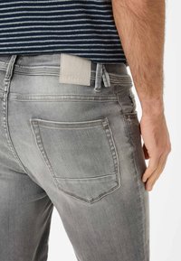 Jeans en denim gris clair avec un design standard à cinq poches, un léger délavage et une étiquette en simili cuir sur la ceinture arrière.