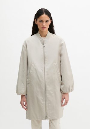 Vrouw die een lange, beige jas met ritssluiting draagt, met opgeblazen mouwen en zijzakken, staand tegen een effen witte achtergrond.